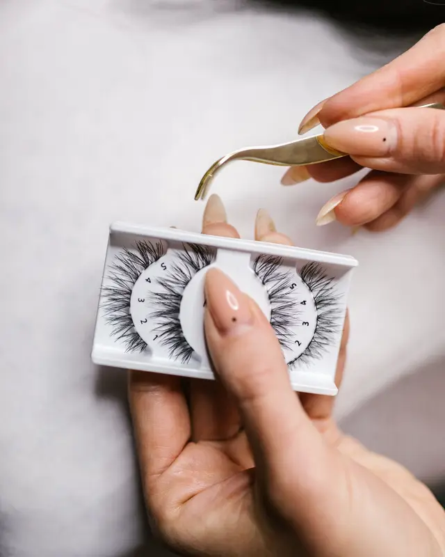 Hands holding gold tweezers over a tray of false lashes, ready to pick one up