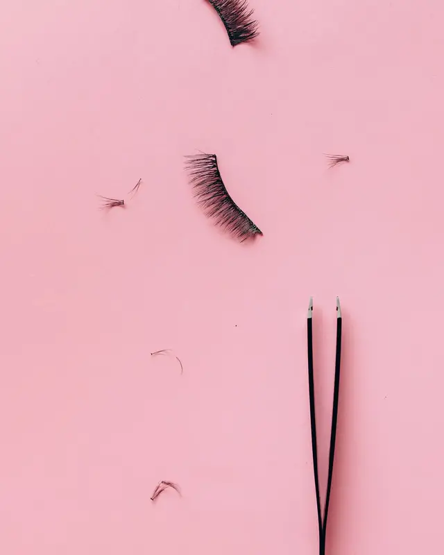 Flat lay of two false eyelash strips, loose lash clusters and black tweezers on a pink background